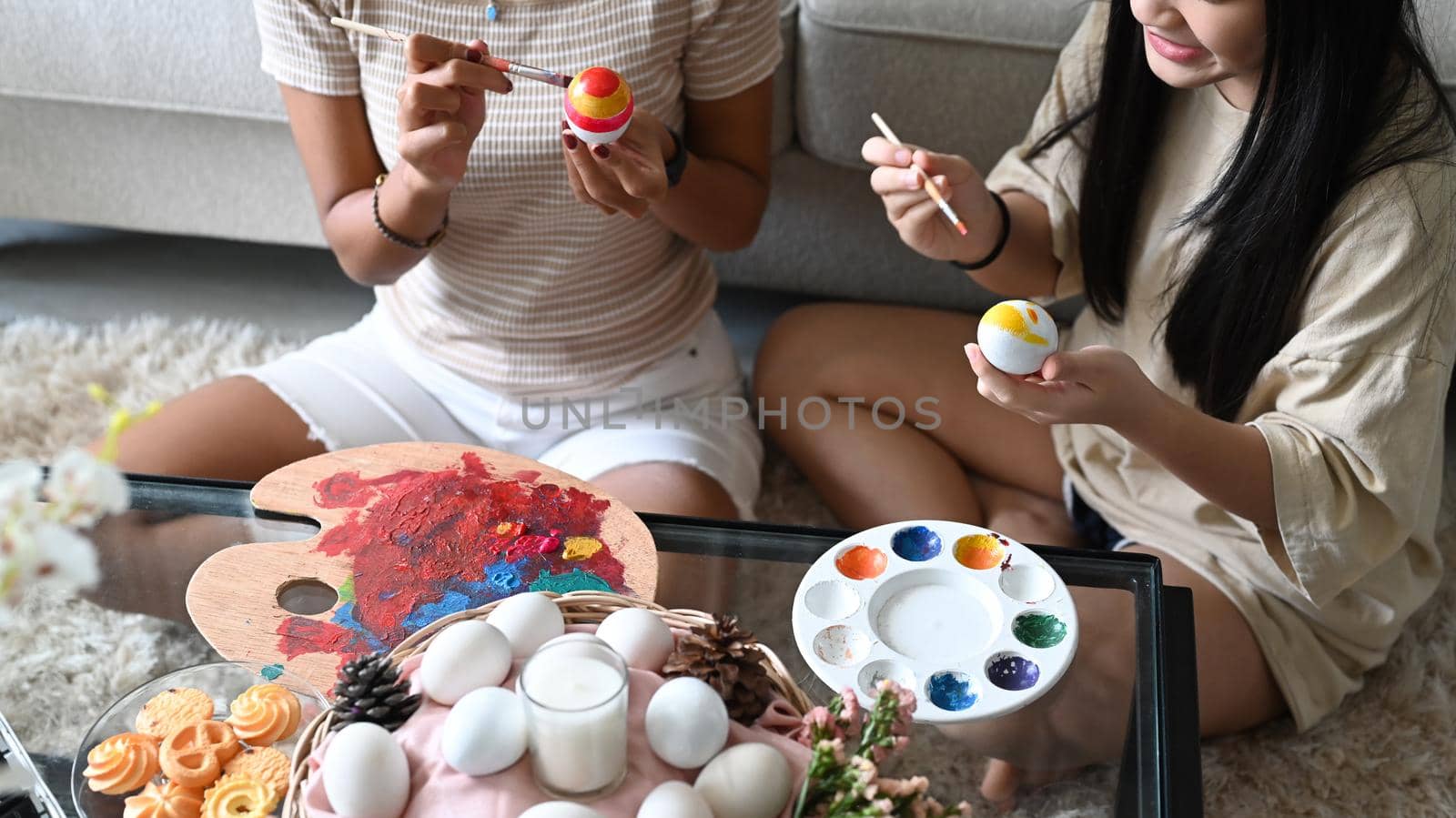 Smiling little girl and mother sitting floor in living room and painting Easter eggs together. Easter, holidays and people concept. by prathanchorruangsak