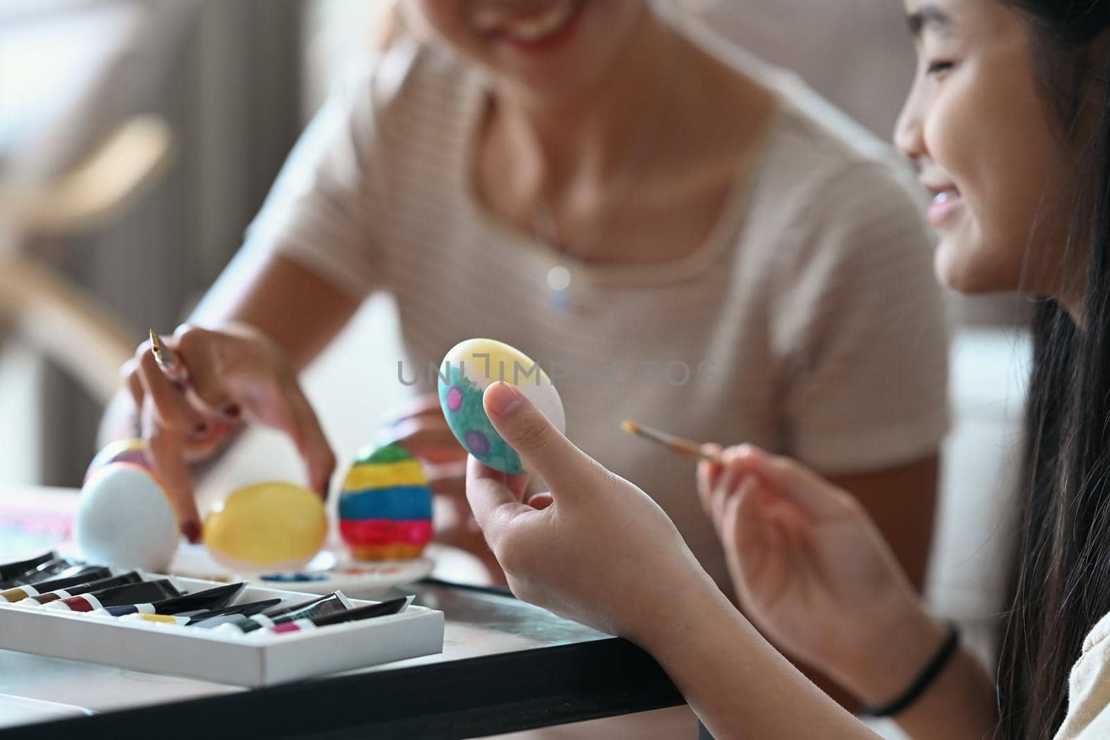 Little girl painting Easter eggs with mother while preparing for Easter holiday at home together. by prathanchorruangsak