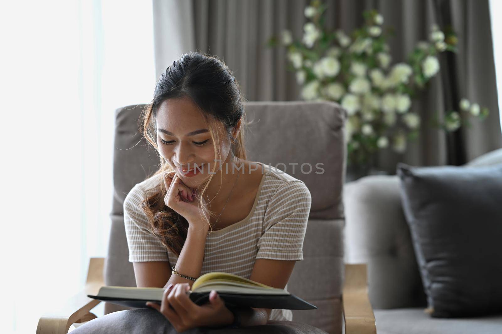 Happy millennial woman reading book while sitting on beige armchair in apartment.