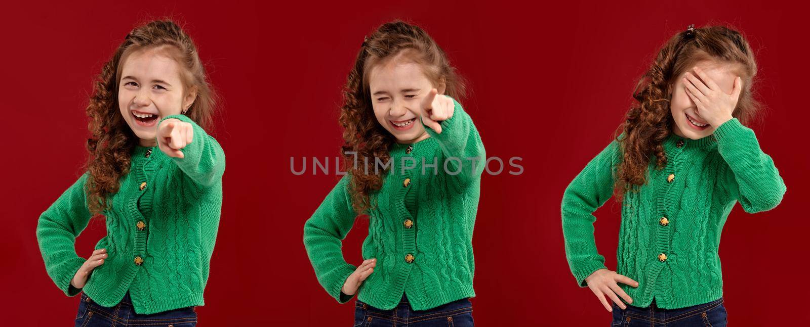 Close-up portrait of a lovely curly kid in a green knitted sweater laughing at someone while posing on a red studio background. Set of people sincere emotions, lifestyle concept. Mockup copy space.