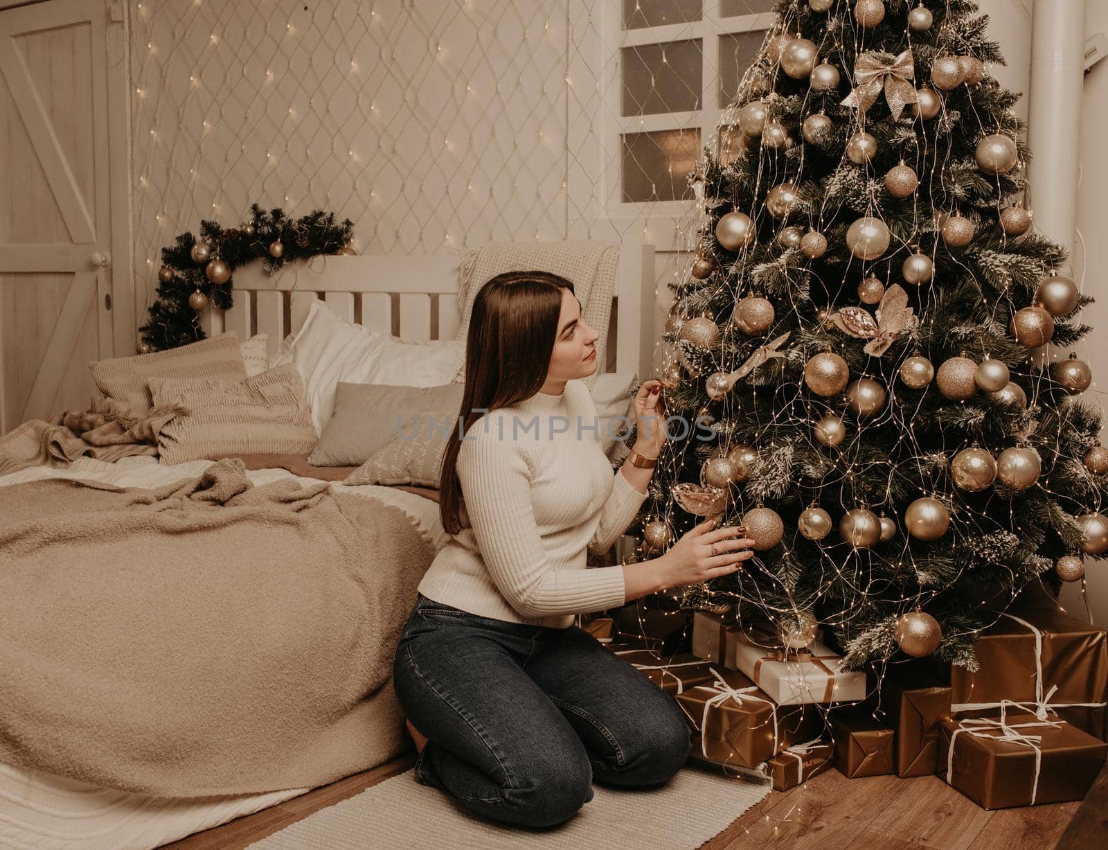 young woman sits in a white golf shirt near Christmas tree in bedroom. decorated house for New Year. Christmas morning. apartment interior. Valentine's Day celebration