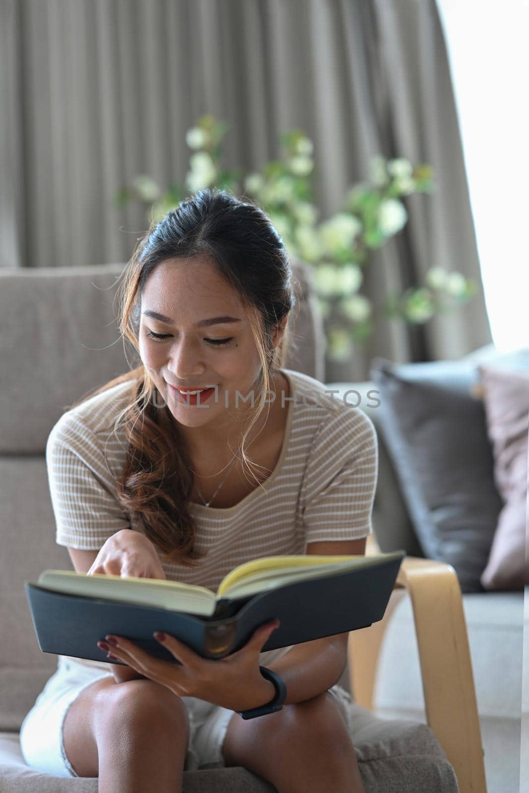 Peaceful young asian woman reading book while resting on comfortable armchair at home.