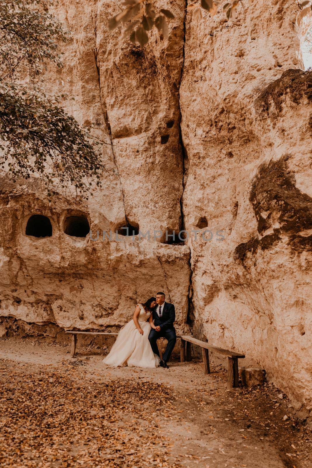 wedding couple in love man and woman sitting on a bench under rock monastery bakota in autumn forest against background of stone rocks. groom suit and bride dress with crown on head