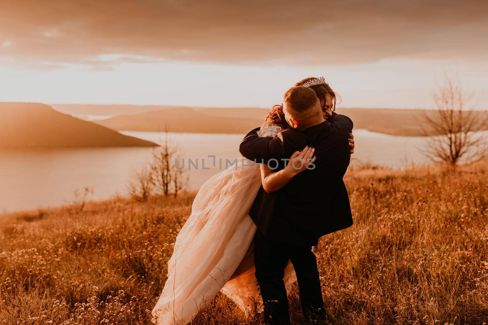 A loving couple wedding newlyweds in a white dress and a suit walk run smile happy on tall grass in the summer field on the mountain above the river. sunset.