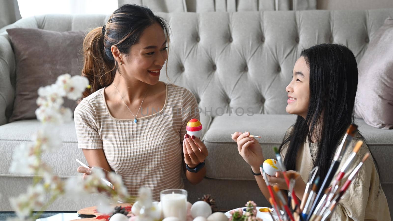 Happy young mother and cute daughter are sitting in bright living room and coloring Easter eggs.