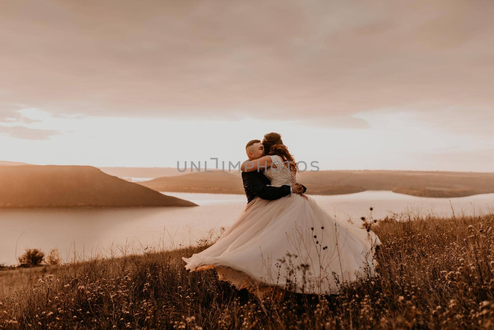 loving couple wedding newlyweds in white dress and suit hug kissing whirl on tall grass in summer field on mountain above the river. sunset.