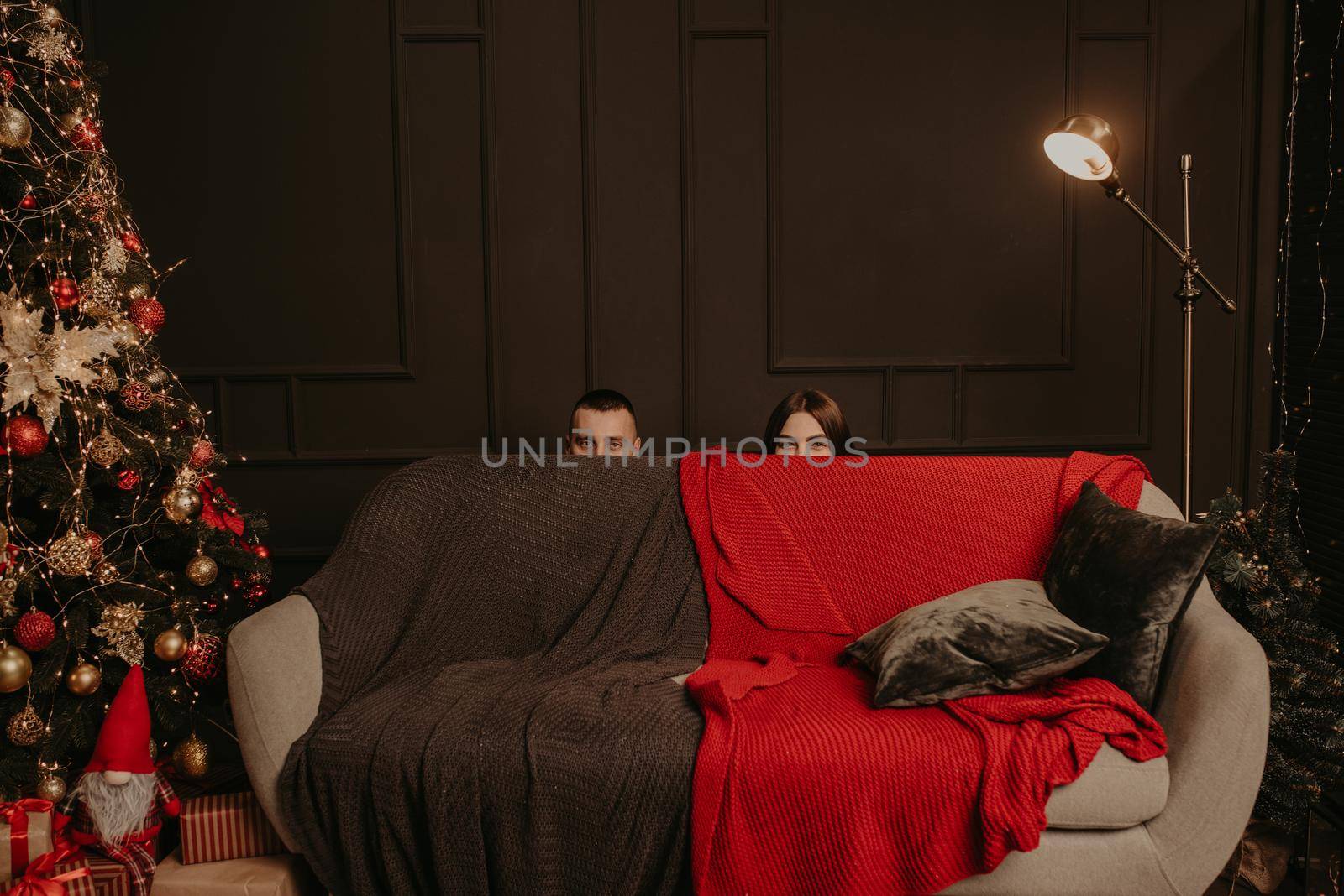 man and a woman hid behind couch. heads of a man and a woman stick out from behind sofa. decorated house for New Year. Christmas morning. apartment interior. Valentine's Day celebration