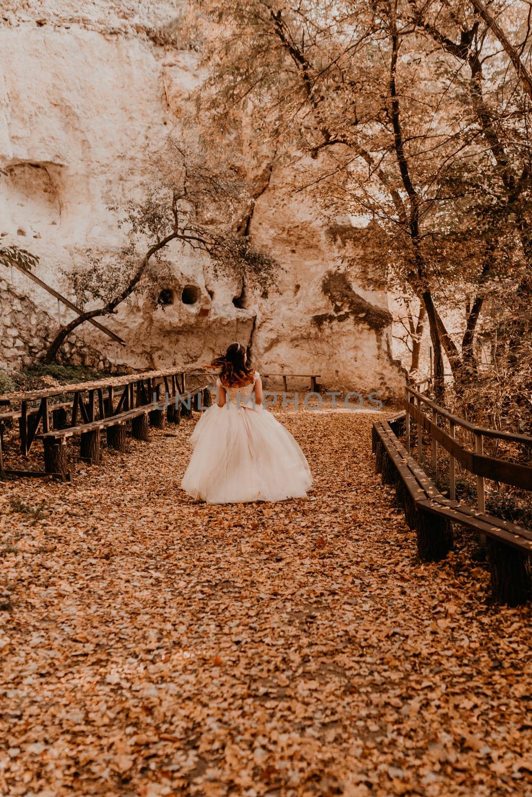 woman bride in white wedding dress with hairstyle makeup and crown on her head walks through autumn forest on fallen orange leaves