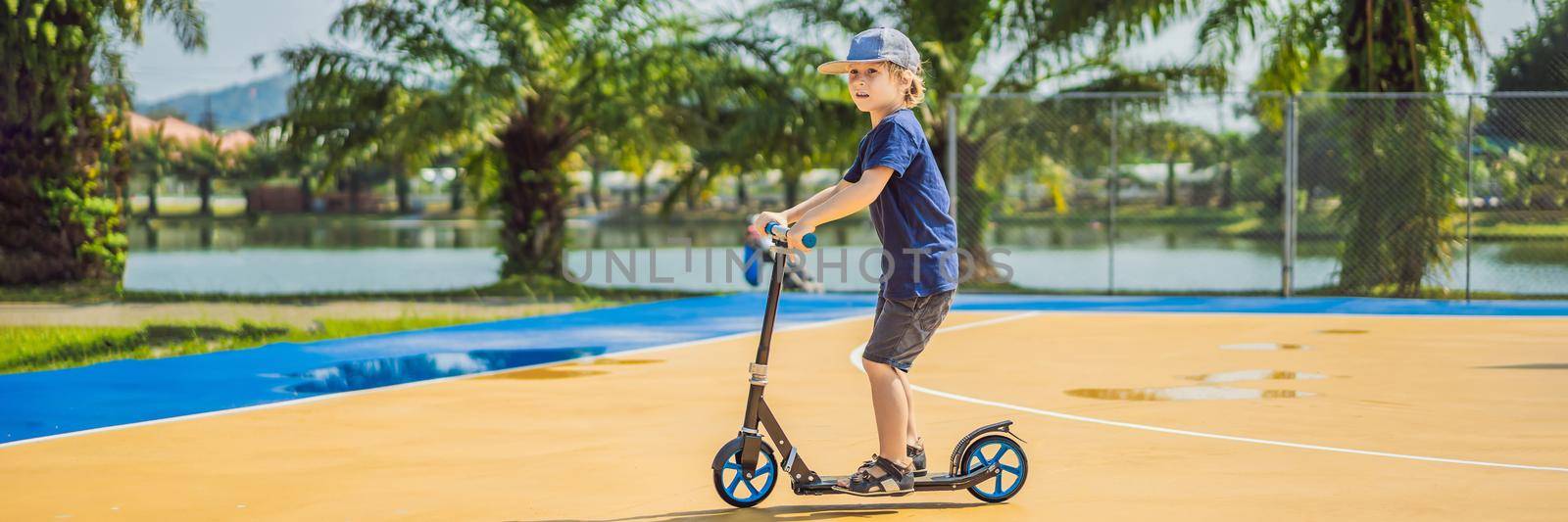 BANNER, LONG FORMAT Happy child on kick scooter in on the basketball court. Kids learn to skate roller board. Little boy skating on sunny summer day. Outdoor activity for children on safe residential street. Active sport for preschool kid.