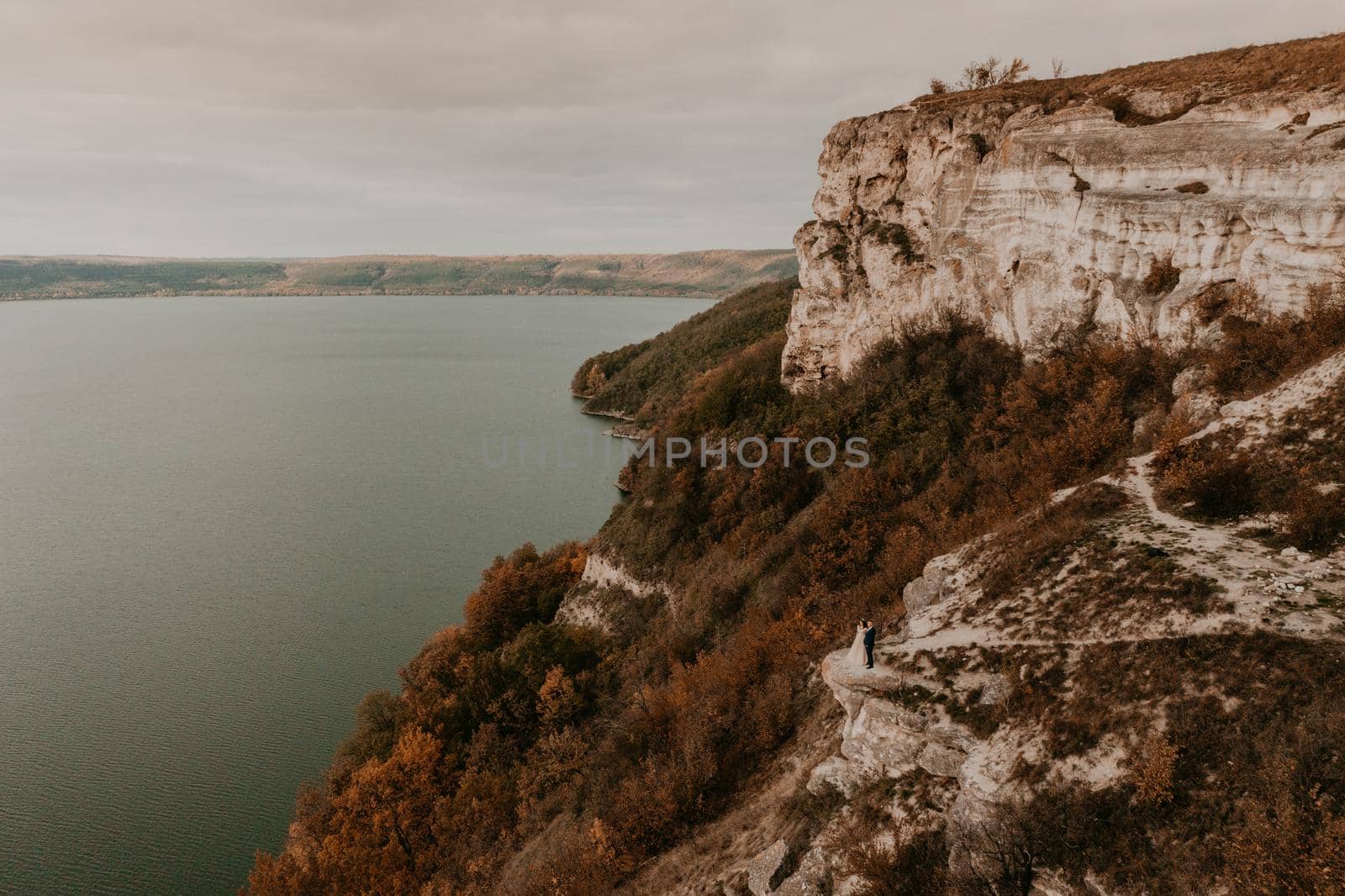 groom in suit and the bride in a dress stand on a stone cliff above the river. Mountain near lake. Aerial view from drone. rock monastery in bakota