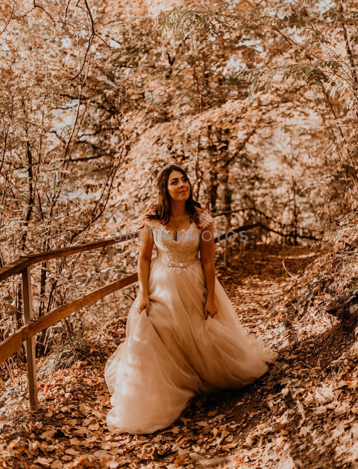 woman bride in white wedding dress with hairstyle makeup and crown on her head walks through autumn forest on fallen orange leaves
