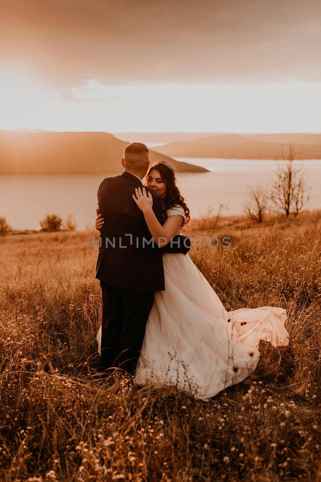A loving couple wedding newlyweds in white dress and suit walk hug kissing whirl on tall grass in summer field on mountain above the river.style bride accessories on head crown makeup hairstyle