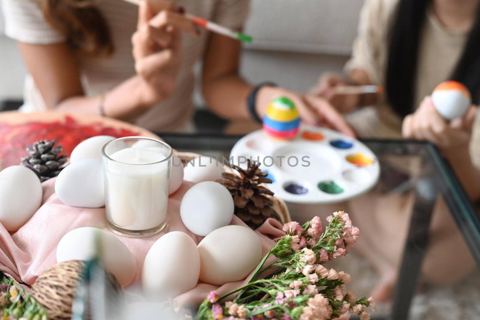 Wicker basket full of eggs and flowers on table in living room with mother and daughter painting Easter egg in background.