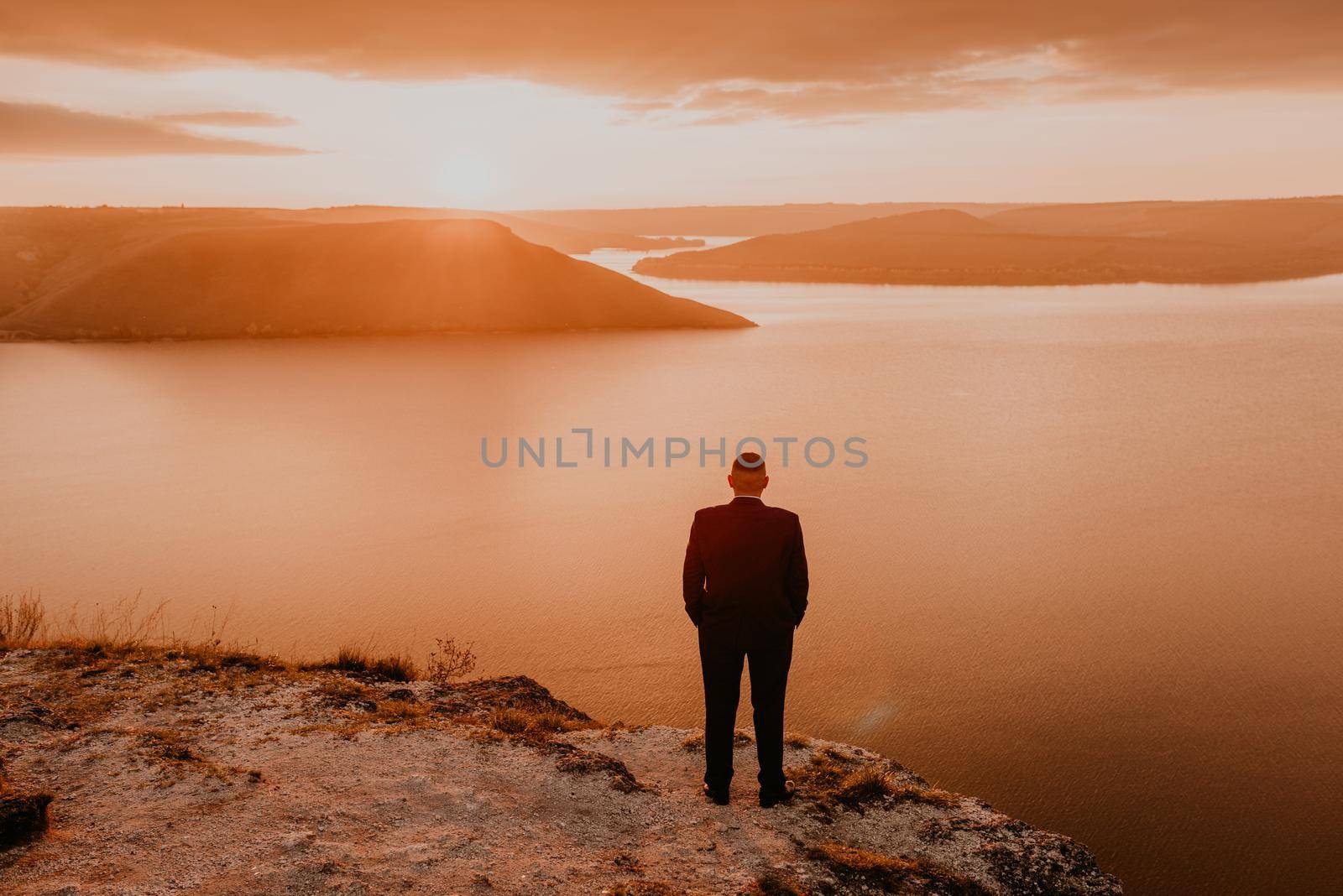 man in formal suit groom stands on mountain holds his hands in pockets. fiery orange sunset sunset rays through clouds. panoramic view from cliff to river lake sea in distance silhouettes islands