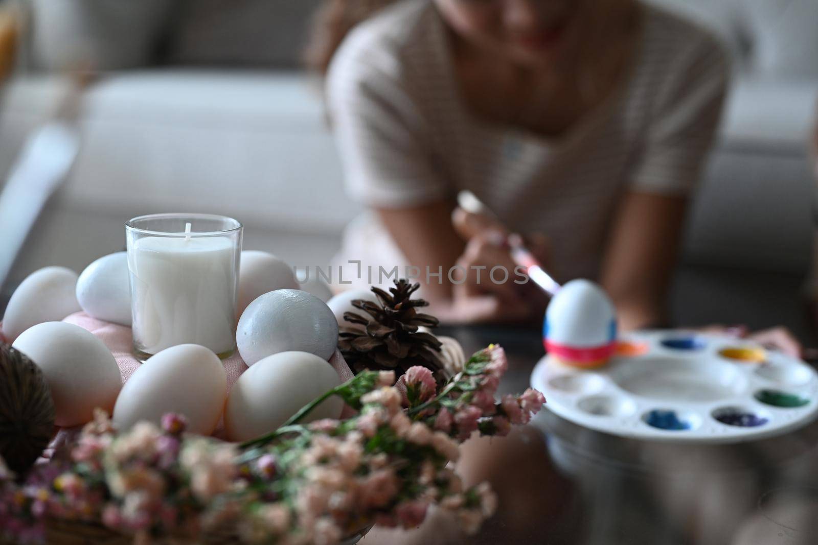 Close up view wicker basket full of eggs, candle and flowers on table with young woman painting Easter egg in background.