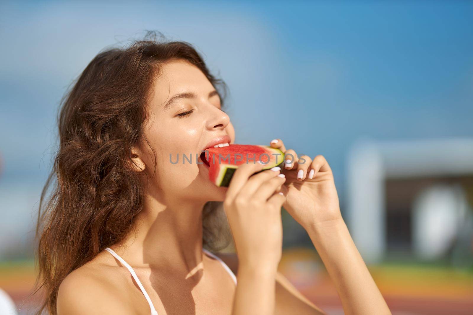 Young brunette woman biting juicy slice of watermelon outdoor. Portrait view of charming caucasian girl eating ripe watermelon, closing eyes in pleasure, with copy space. Concept of satisfaction.