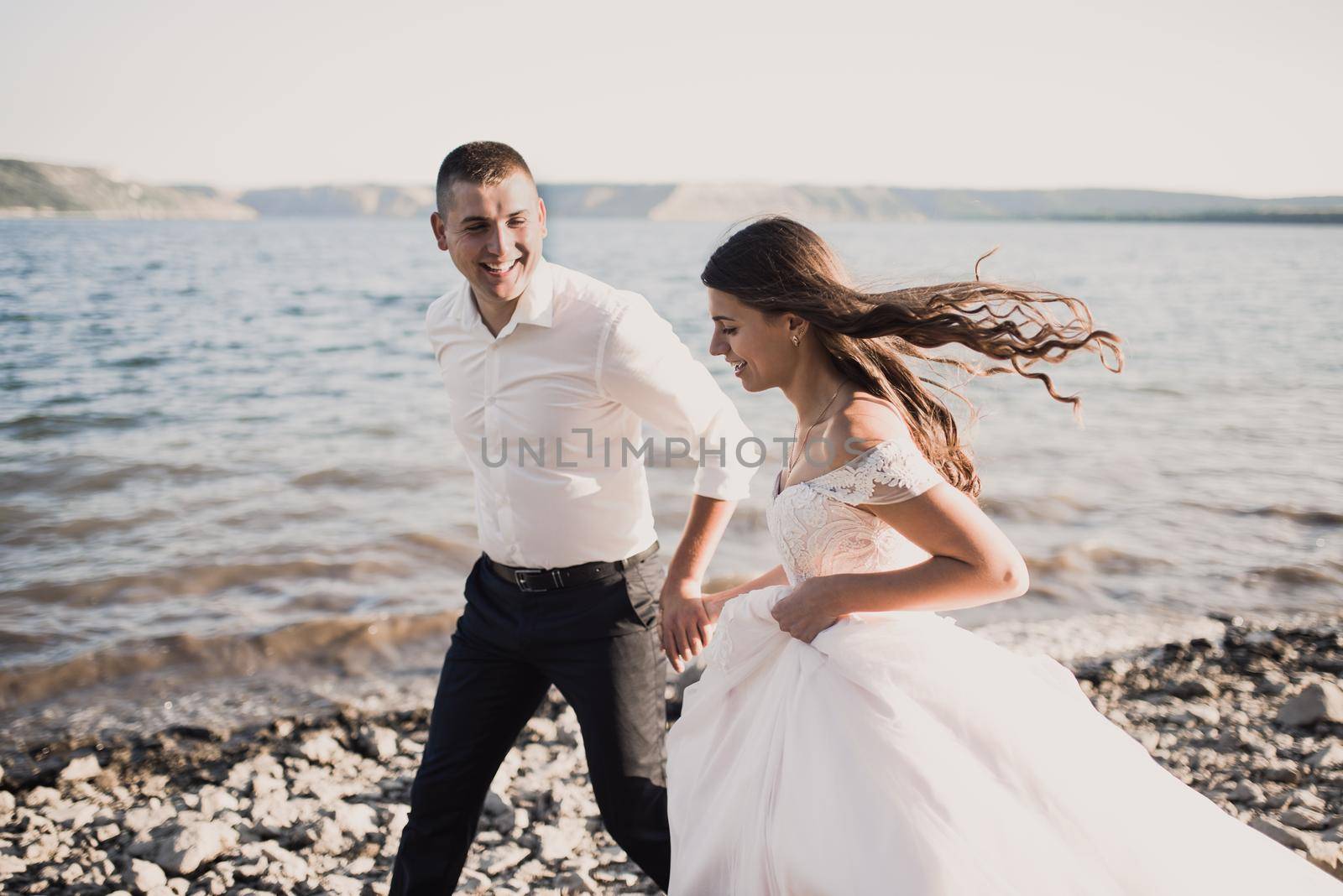 bride and groom laughing run together against rays of setting sun in summer by AndriiDrachuk