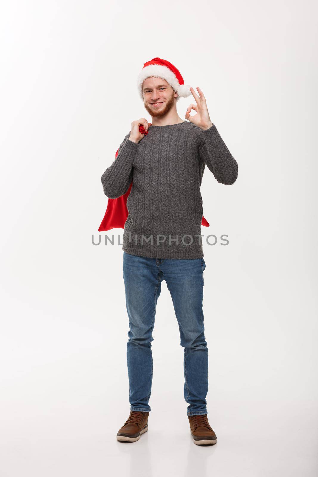 Christmas concept - Young confident smart man holding red big santa bag and giving ok sign.