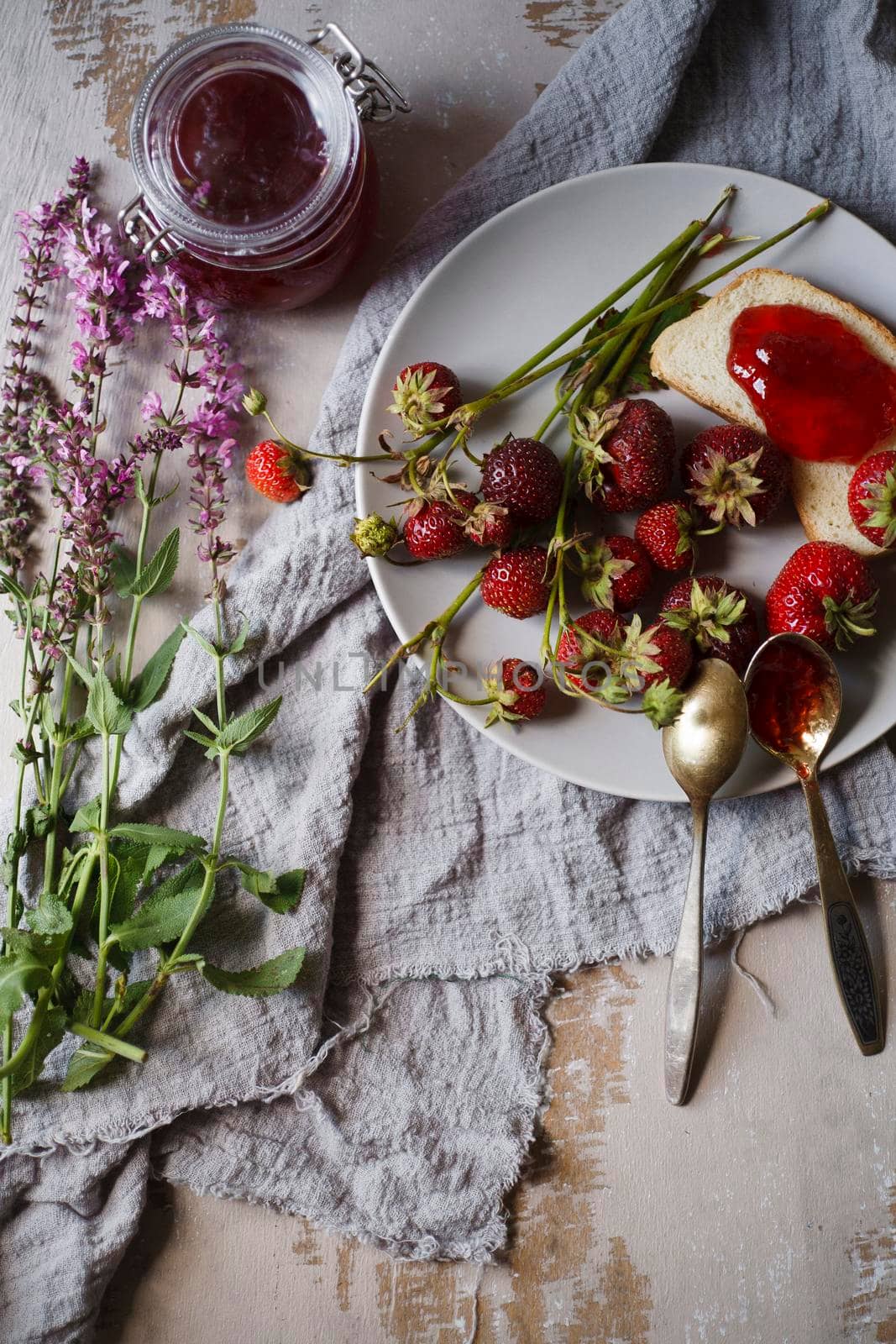 Summer breakfast concept with srawberry jam with bread and fresh harvested berries on wooden rustic table with pink flowers, top view, flat lay, selective focus.