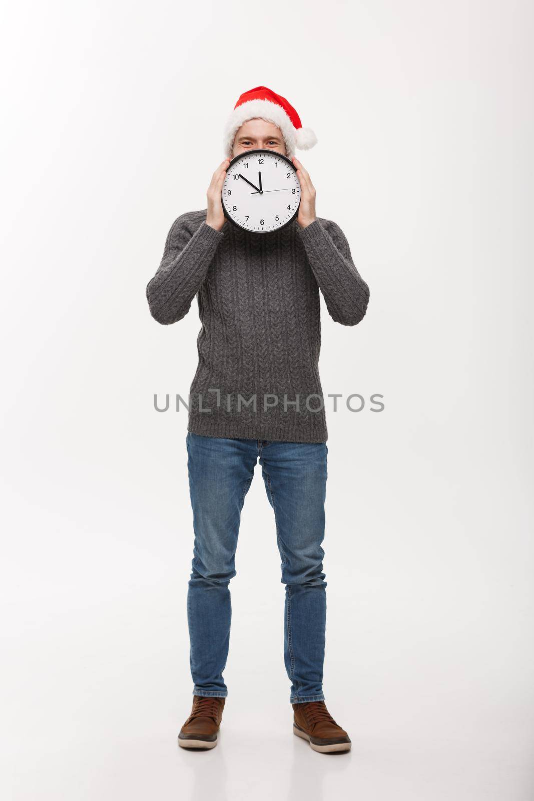 Holiday concept - Young handsome beard man hidden behind white clock over white studio background.