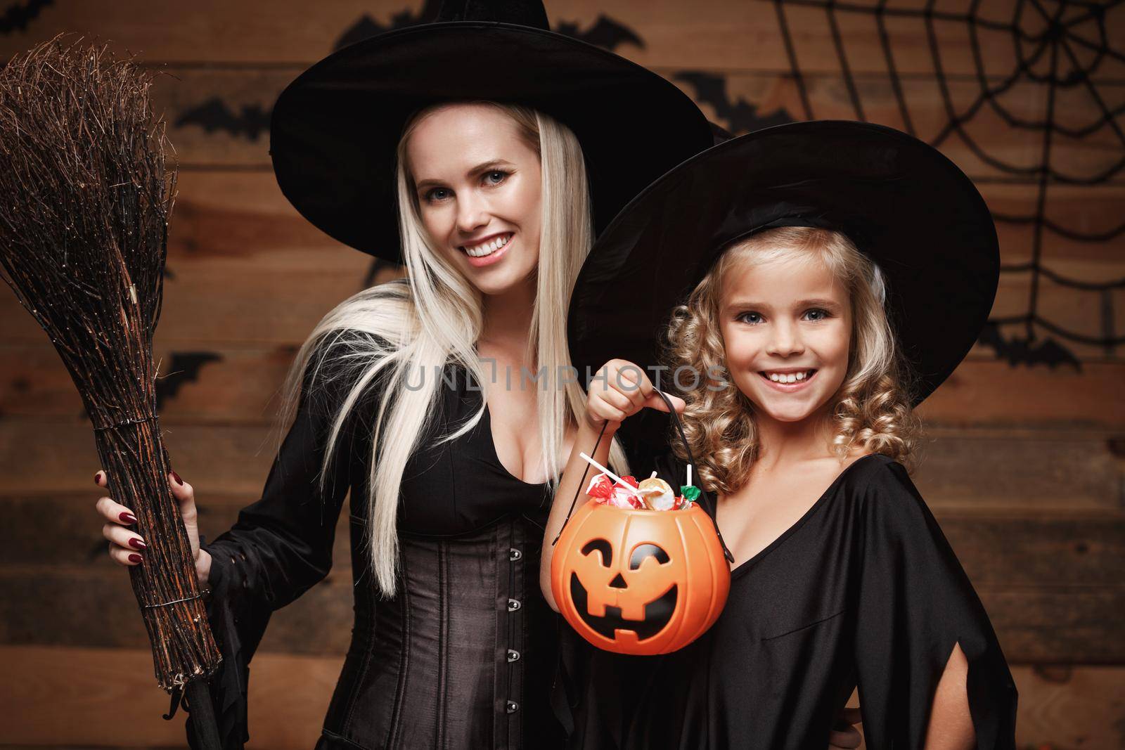 Halloween Concept: beautiful caucasian mother and her daughter in witch costumes celebrating Halloween with sharing Halloween candy and sweet over bats and spider web on Wooden studio background.