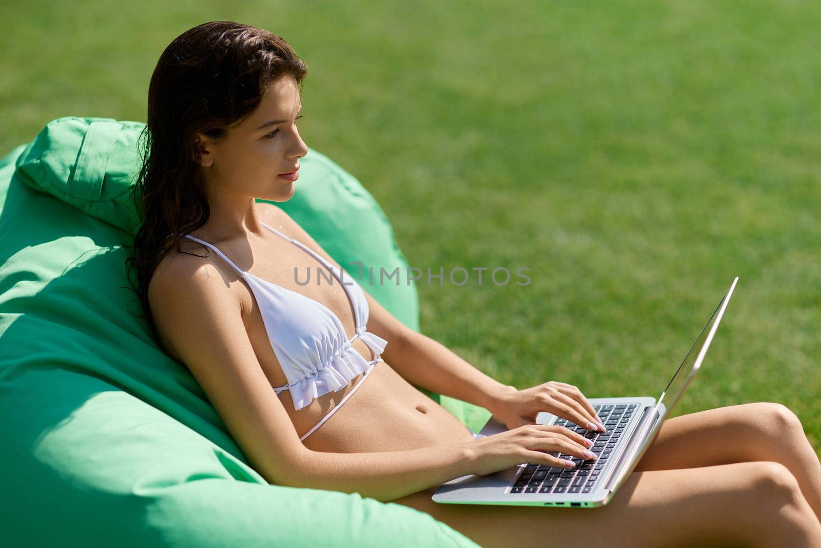Attractive brunette girl sitting on green pillow, while surfing internet on vacation. Side view of slim female freelancer typing on laptop, while resting on bag chair at resort. Concept of freelance.