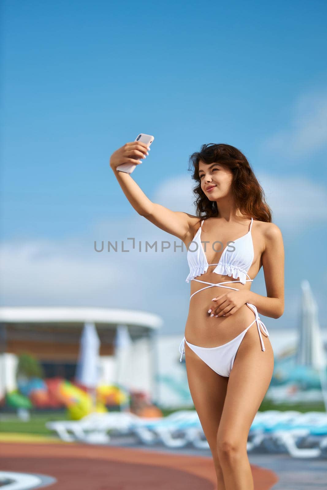 Young smiling woman wearing white swimsuit sunbathing on pool edge while taking photo in summer. Front view of pretty brunette girl taking selfie with mobile phone at resort. Concept of lifestyle.