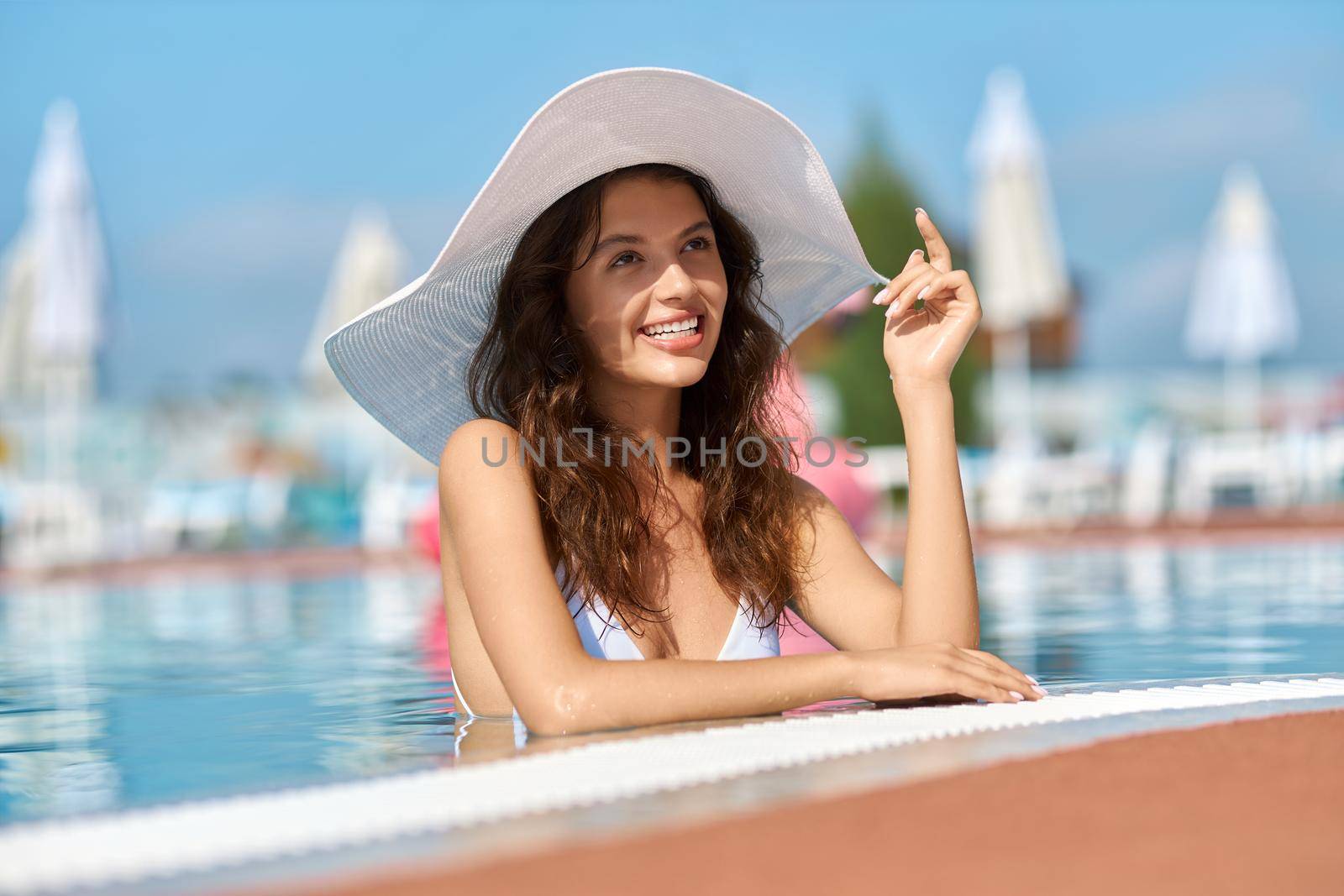 Smiling happy woman wearing trendy white hat standing in swimming pool outdoors. by SerhiiBobyk