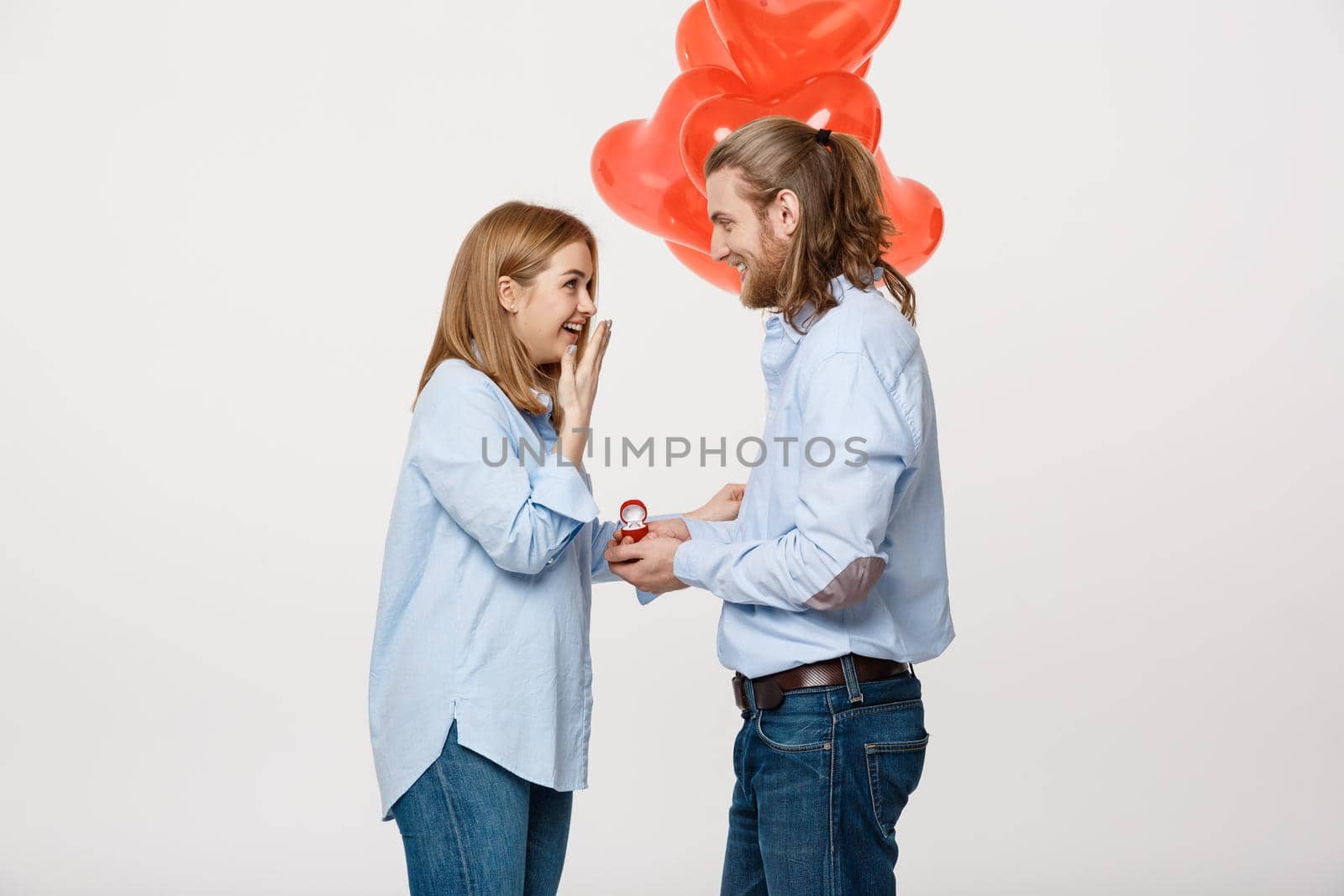 Portrait of Young handsome guy gives a ring to a girl on a white background with red heart air balloons. by Benzoix