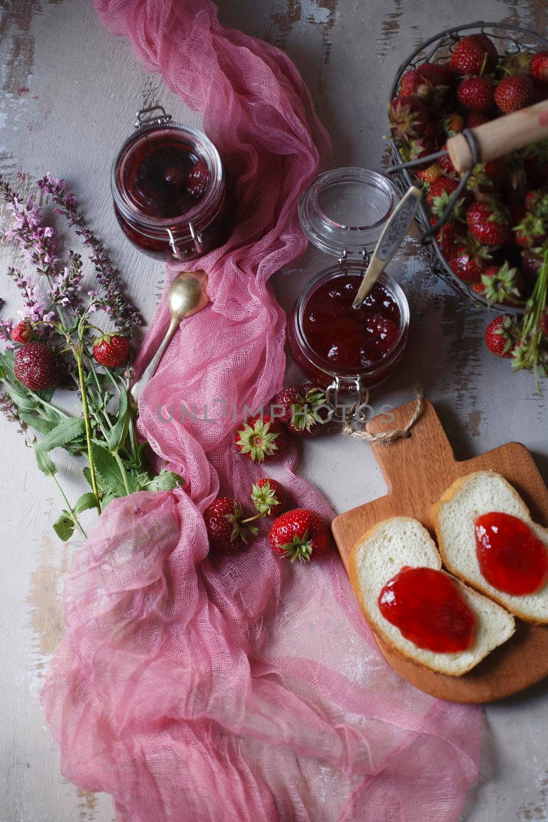 Summer breakfast concept with srawberry jam with bread and fresh harvested berries in basket with pink flowers and cloth, top view, flat lay, selective focus.