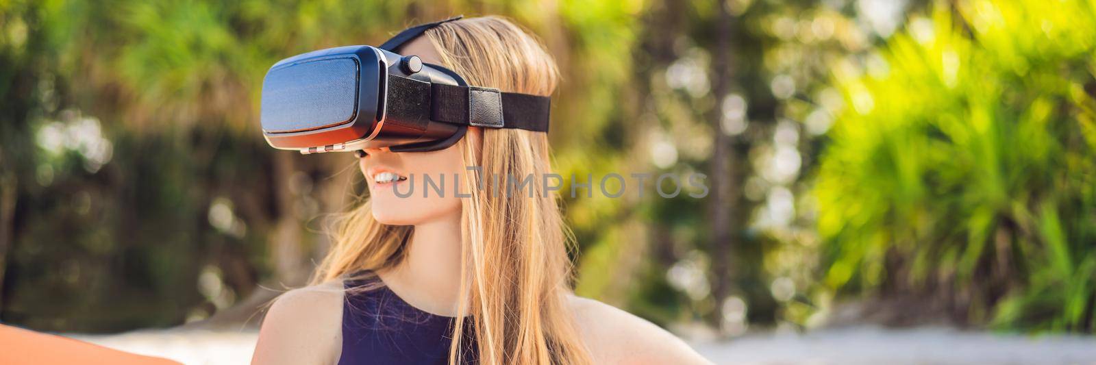 BANNER, LONG FORMAT Summer lifestyle portrait of pretty girl sitting on the orange inflatable sofa and uses virtual reality headset on the beach of tropical island. Relaxing and enjoying life on air bed by galitskaya