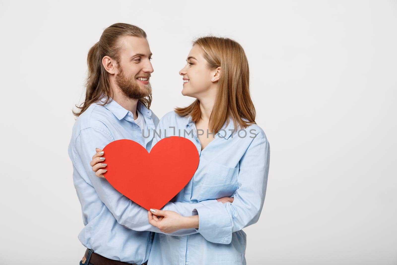 Portrait of happy cute couple in love enjoys Valentine's Day. A man with a beard and a woman with blond short hair holding red heart paper. by Benzoix
