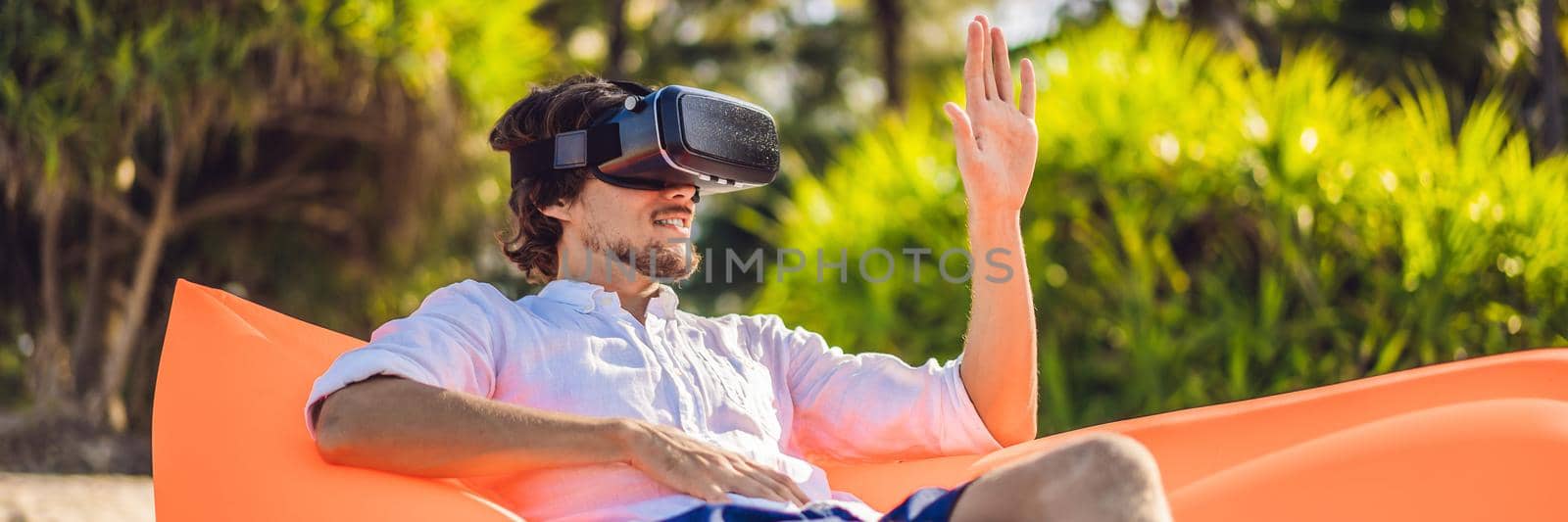 BANNER, LONG FORMAT Summer lifestyle portrait of man sitting on the orange inflatable sofa and uses virtual reality headset on the beach of tropical island. Relaxing and enjoying life on air bed.