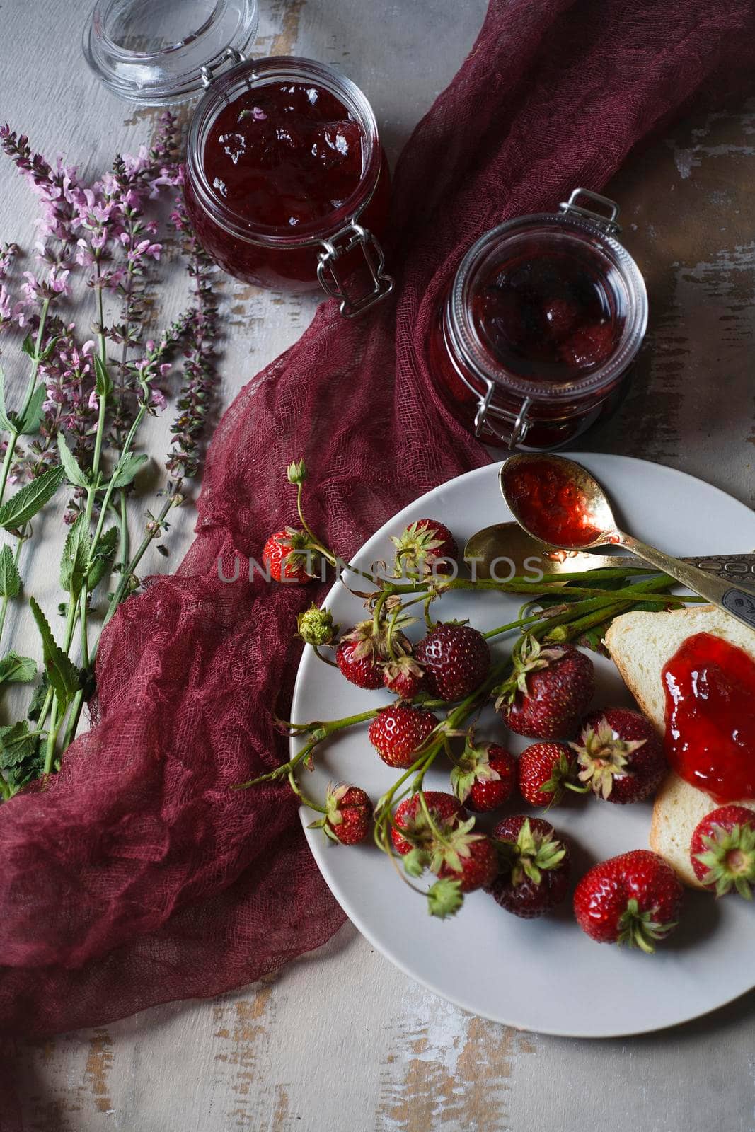 Summer breakfast concept with srawberry jam with bread and fresh harvested berries on wooden rustic table with pink flowers, top view, flat lay, selective focus.