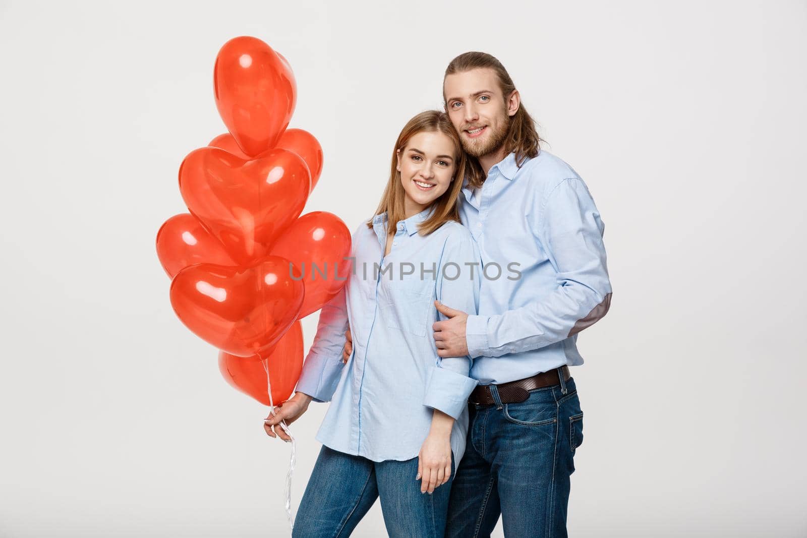 Portrait of young fashionable caucasian couple with balloons heart hugging at each other over isolated white background .