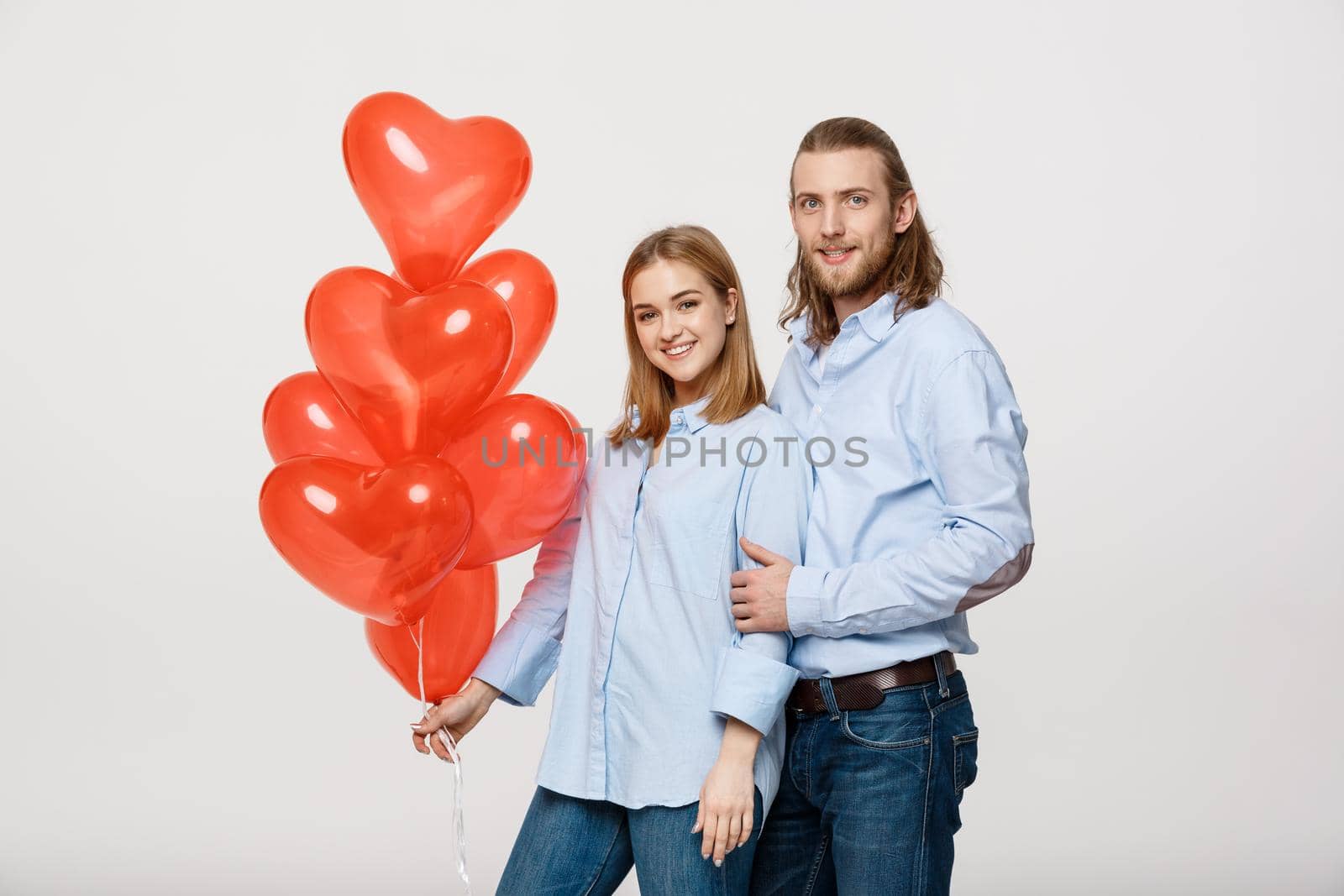 Portrait of young fashionable caucasian couple with balloons heart hugging at each other over isolated white background .