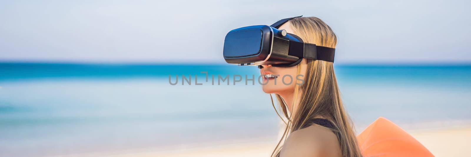 BANNER, LONG FORMAT Summer lifestyle portrait of pretty girl sitting on the orange inflatable sofa and uses virtual reality headset on the beach of tropical island. Relaxing and enjoying life on air bed by galitskaya