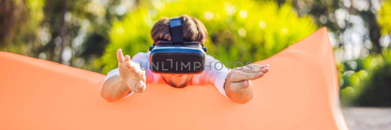 BANNER, LONG FORMAT Summer lifestyle portrait of man sitting on the orange inflatable sofa and uses virtual reality headset on the beach of tropical island. Relaxing and enjoying life on air bed by galitskaya