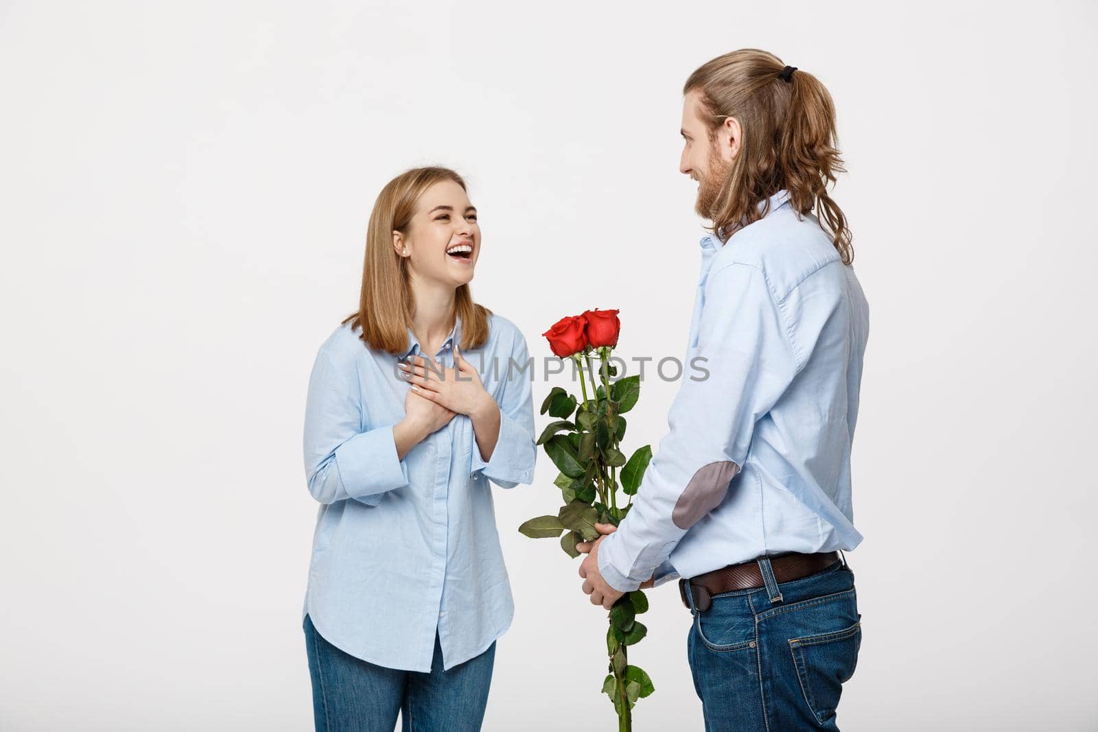 Portrait of handsome elegant guy is surprising his beautiful girlfriend with red roses and smiling over white isolated background .