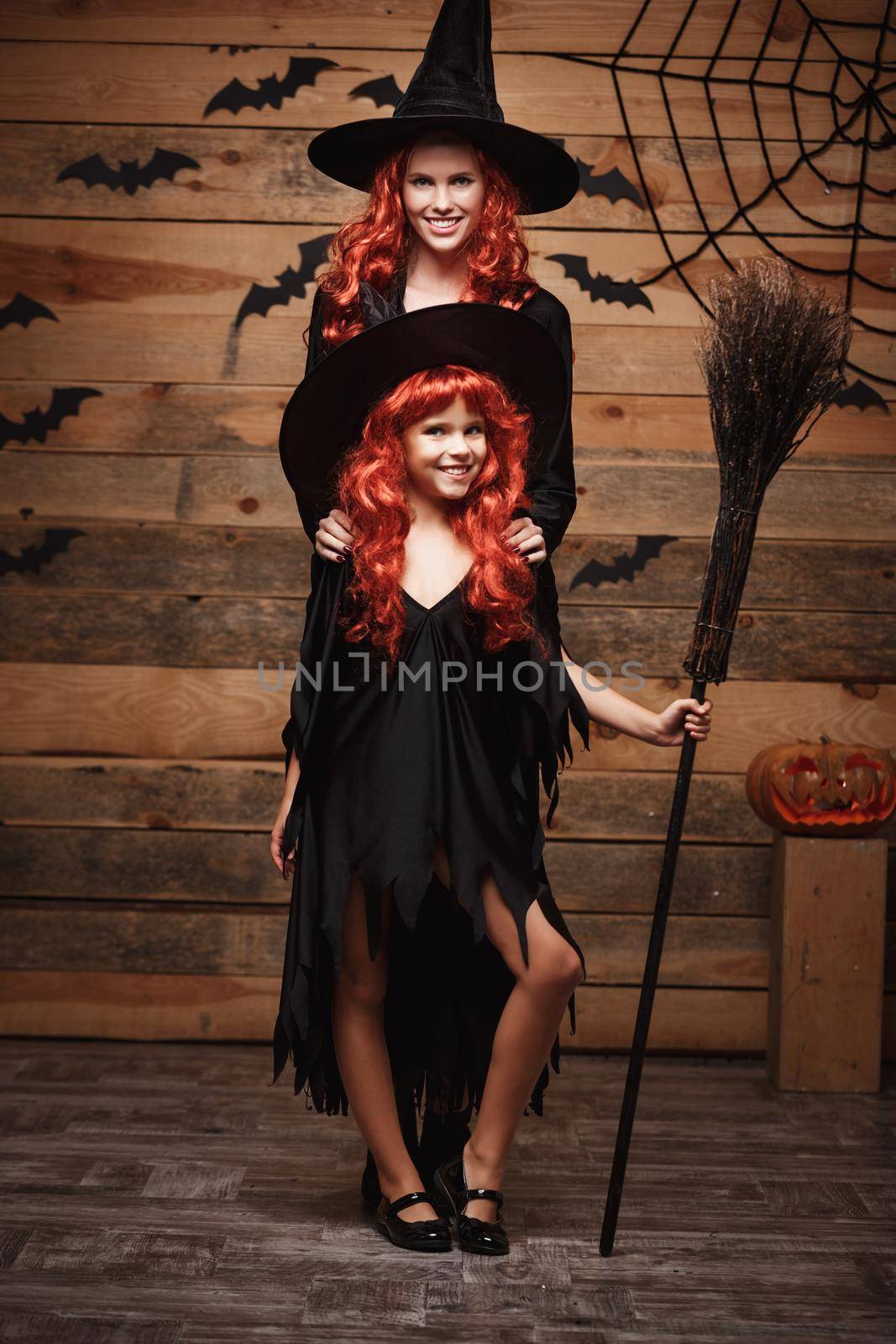 Halloween Concept: Beautiful caucasian mother and her daughter with long red hair in witch costumes celebrating Halloween posing with over bats and spider web on Wooden studio background