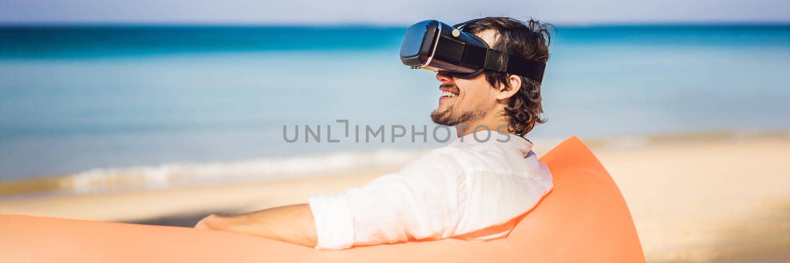 BANNER, LONG FORMAT Summer lifestyle portrait of man sitting on the orange inflatable sofa and uses virtual reality headset on the beach of tropical island. Relaxing and enjoying life on air bed.
