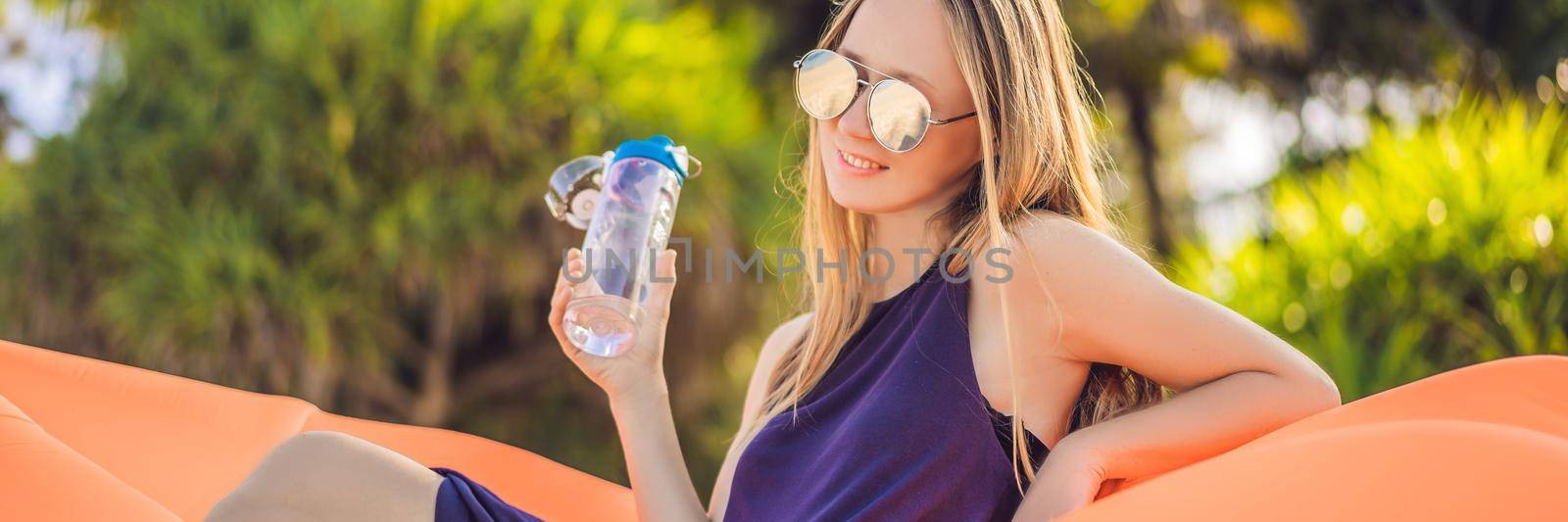 BANNER, LONG FORMAT Summer lifestyle portrait of pretty girl sitting on the orange inflatable sofa and drinking water on the beach of tropical island. Relaxing and enjoying life on air bed.