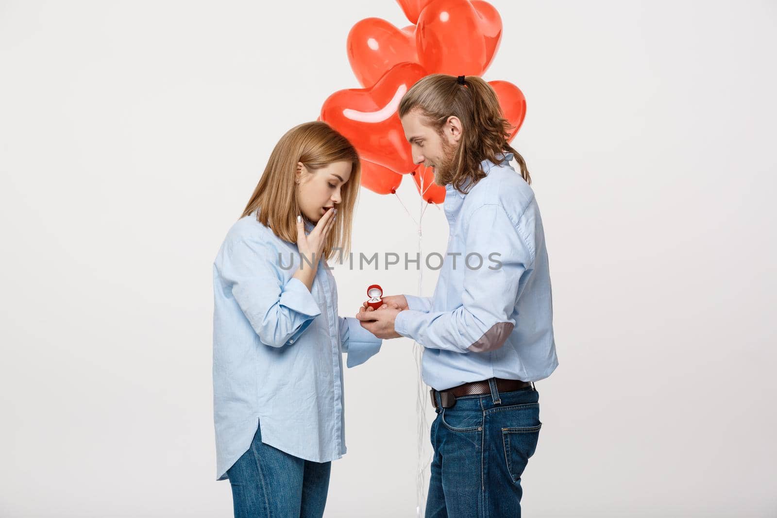 Portrait of Young handsome guy gives a ring to a girl on a white background with red heart air balloons .