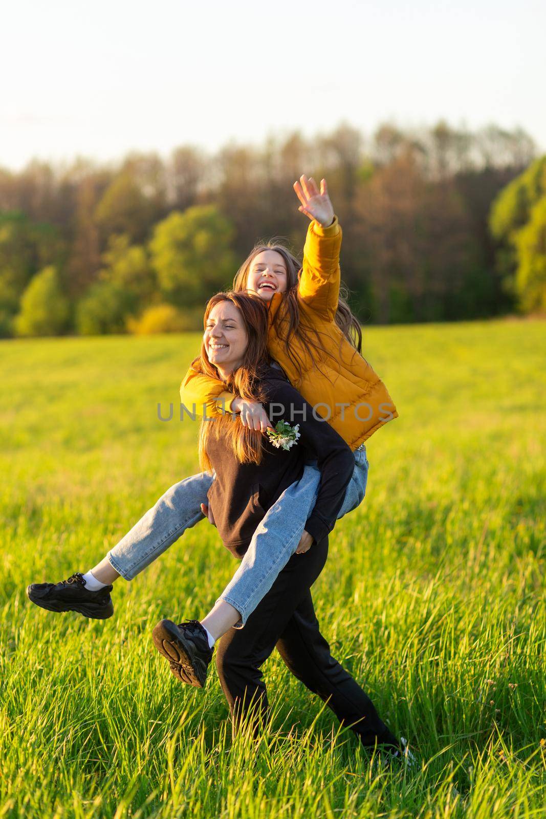 Playful mother giving daughter piggy back ride at green field. by BY-_-BY