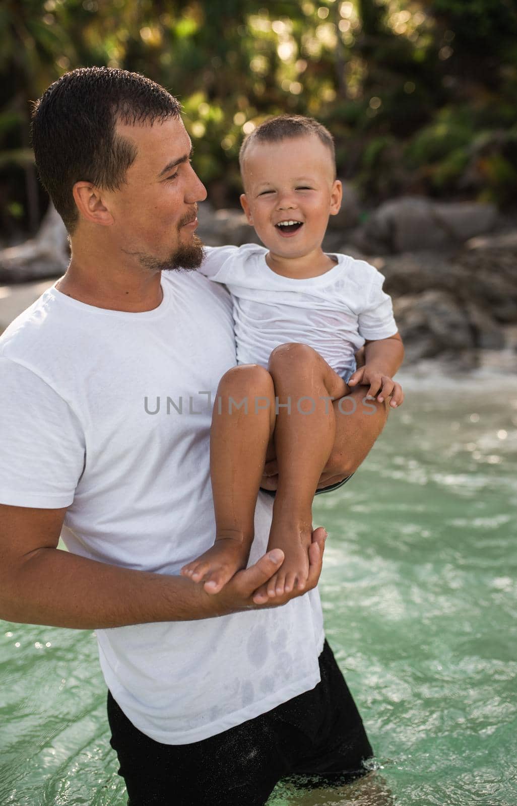 A black-haired young bearded father holds in arms his little child with a blond son.The kid laughs in father arms. family dressed in simple white comfortable clothes.Thailand. Aquamarine crystal sea