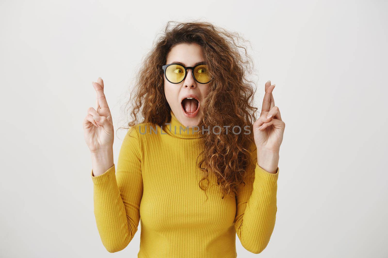 Excited woman in yellow sweater keeping fingers crossed, mouth wide open, waiting for special moment isolated on grey background