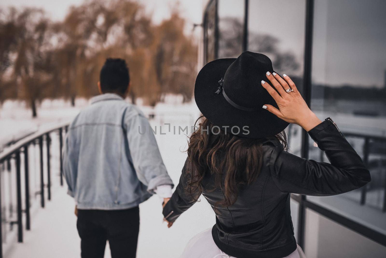 A man in a light blue denim jacket is leading his black-haired curly-haired bride in a white dress with a black leather jacket and a wide-brimmed black hat. Snow lies near the mirror showcases.