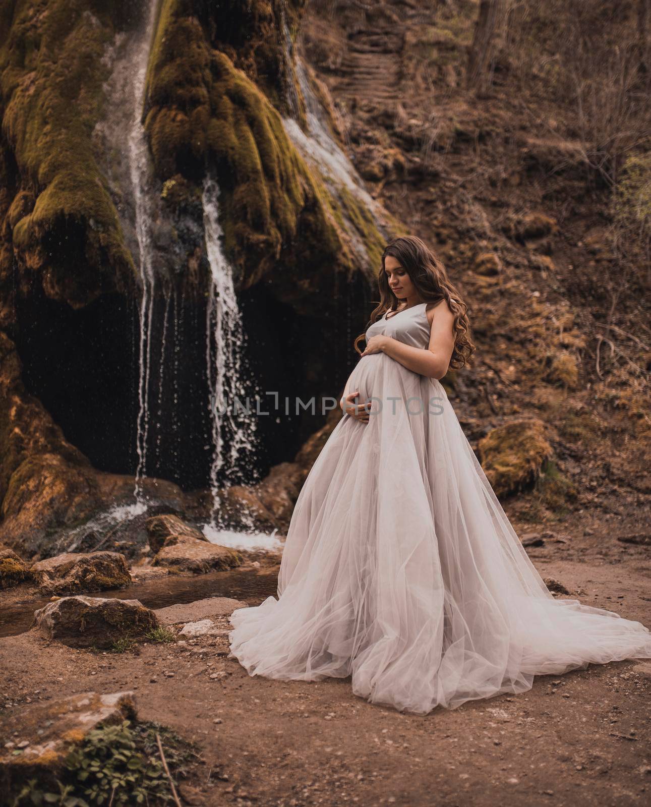 pregnant woman with a big belly in a white long dress in nature on the background of a waterfall