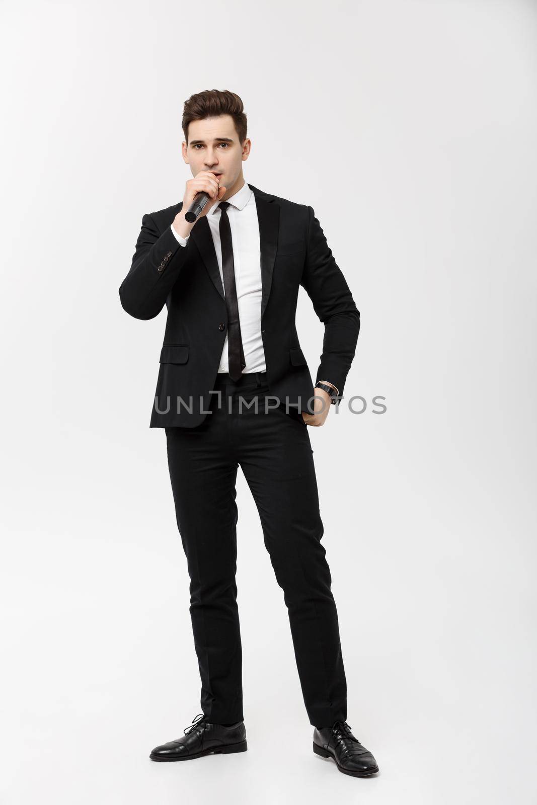 Business Concept: Full-length Portrait young man in black suit is holding a microphone, singing and posing against a white background.