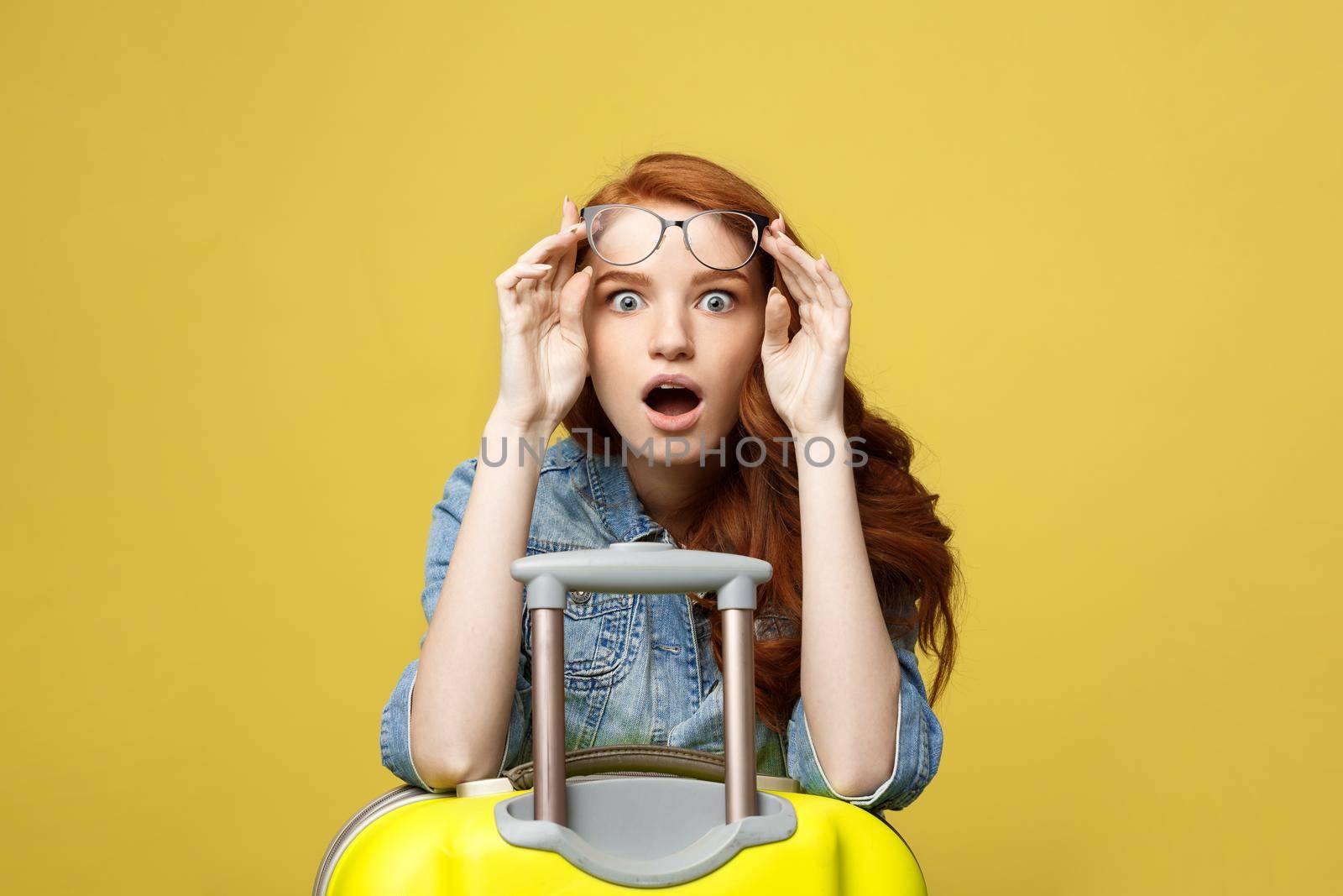 Travel and Lifestyle Concept: Portrait of a shocked girl in denim dress with suitcase looking at camera isolated over golden yellow background.